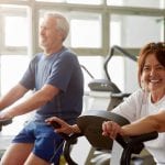 Happy senior woman working out in gym. Smiling elderly couple exercising in gym on stationary bicycle, focus on woman.