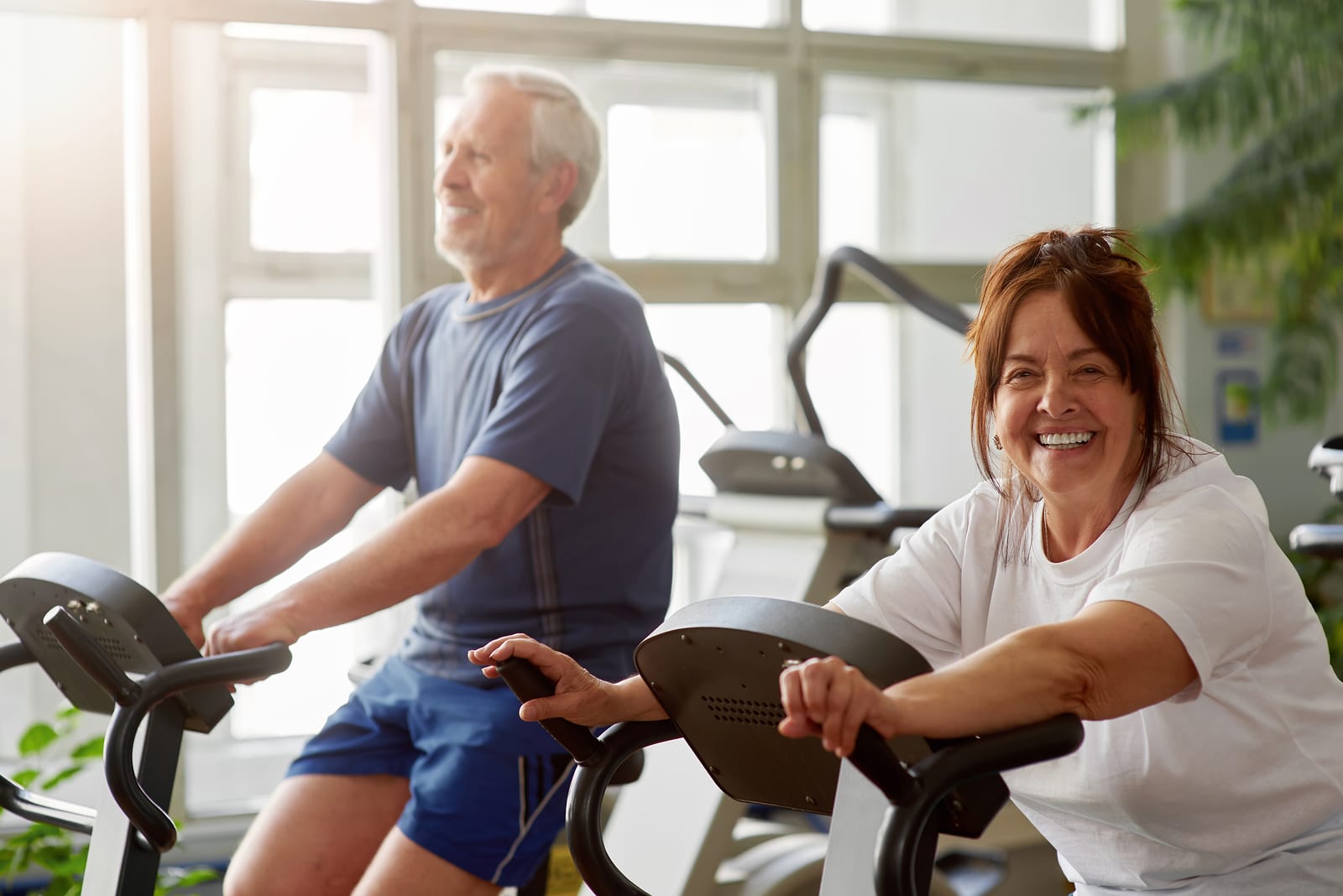 Happy senior woman working out in gym. Smiling elderly couple exercising in gym on stationary bicycle, focus on woman.