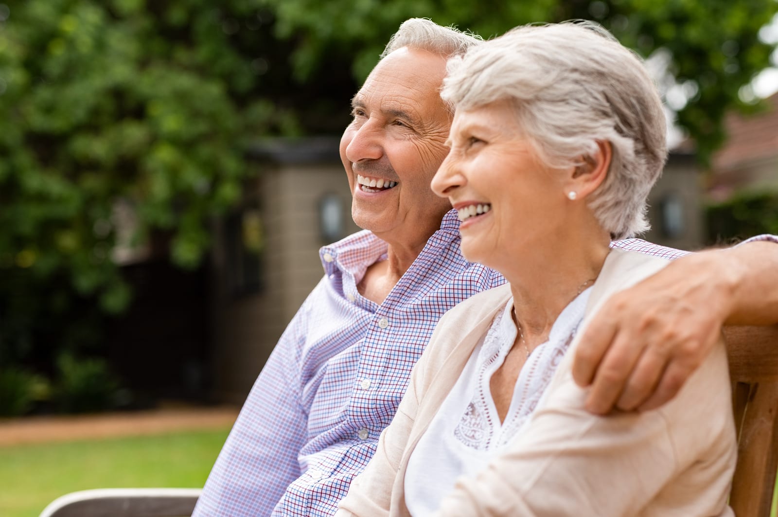 Senior couple sitting together on bench at park. Elderly married couple sitting outdoor and relaxing. Romantic husband embrace his wife while looking away and smiling. Future and retirement concept.