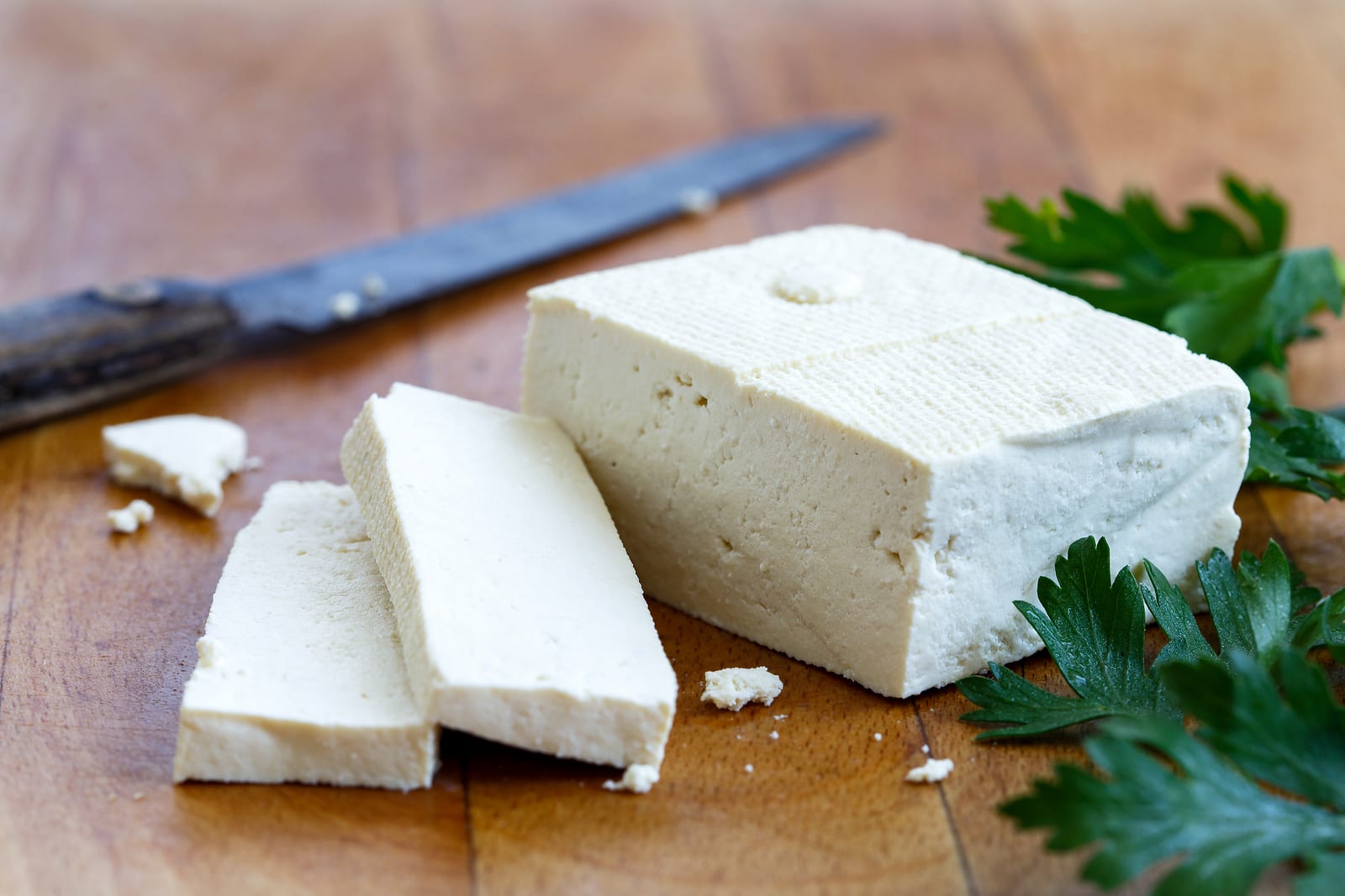 Single block of white tofu with two tofu slices crumbs fresh parsley and rustic knife on wooden chopping board.