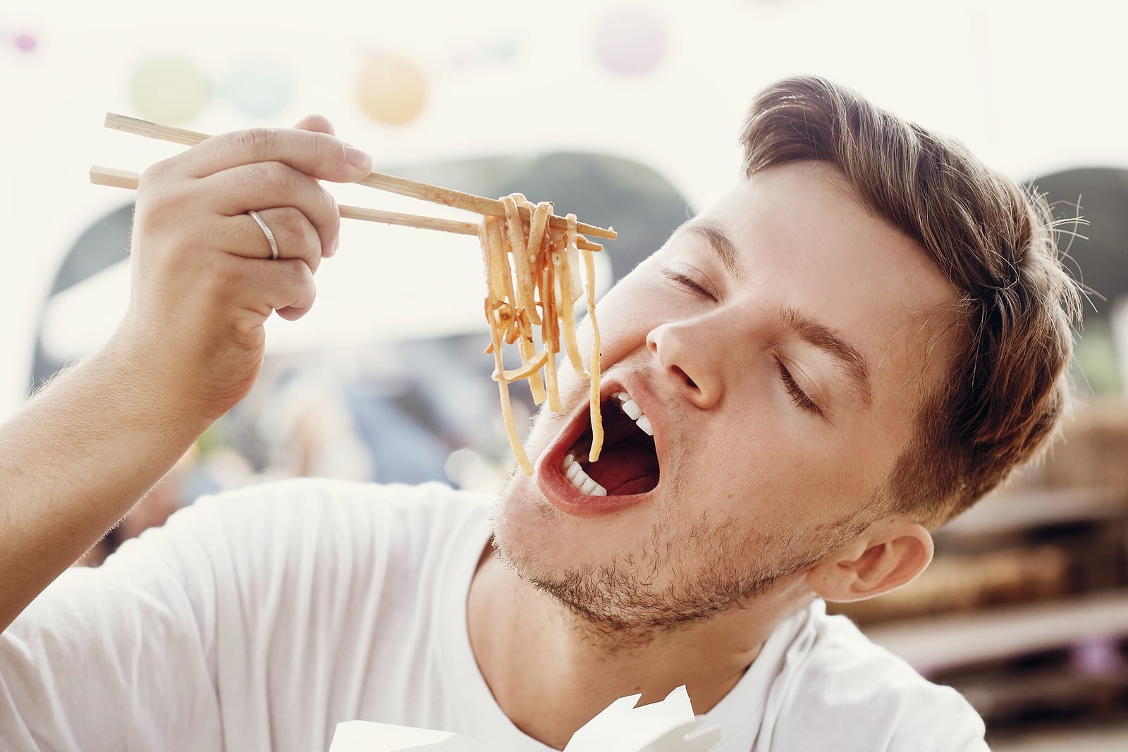 Stylish hungry man eating delicious wok noodles with funny emotions from carton box with bamboo chopsticks. Asian Street food festival. Hipster tasting noodles in takeaway paper box