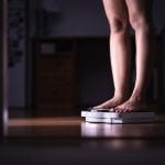 A middle aged woman stands on a scale in a dark hallway. She is checking her weight.