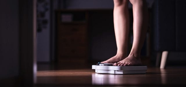 A middle aged woman stands on a scale in a dark hallway. She is checking her weight.