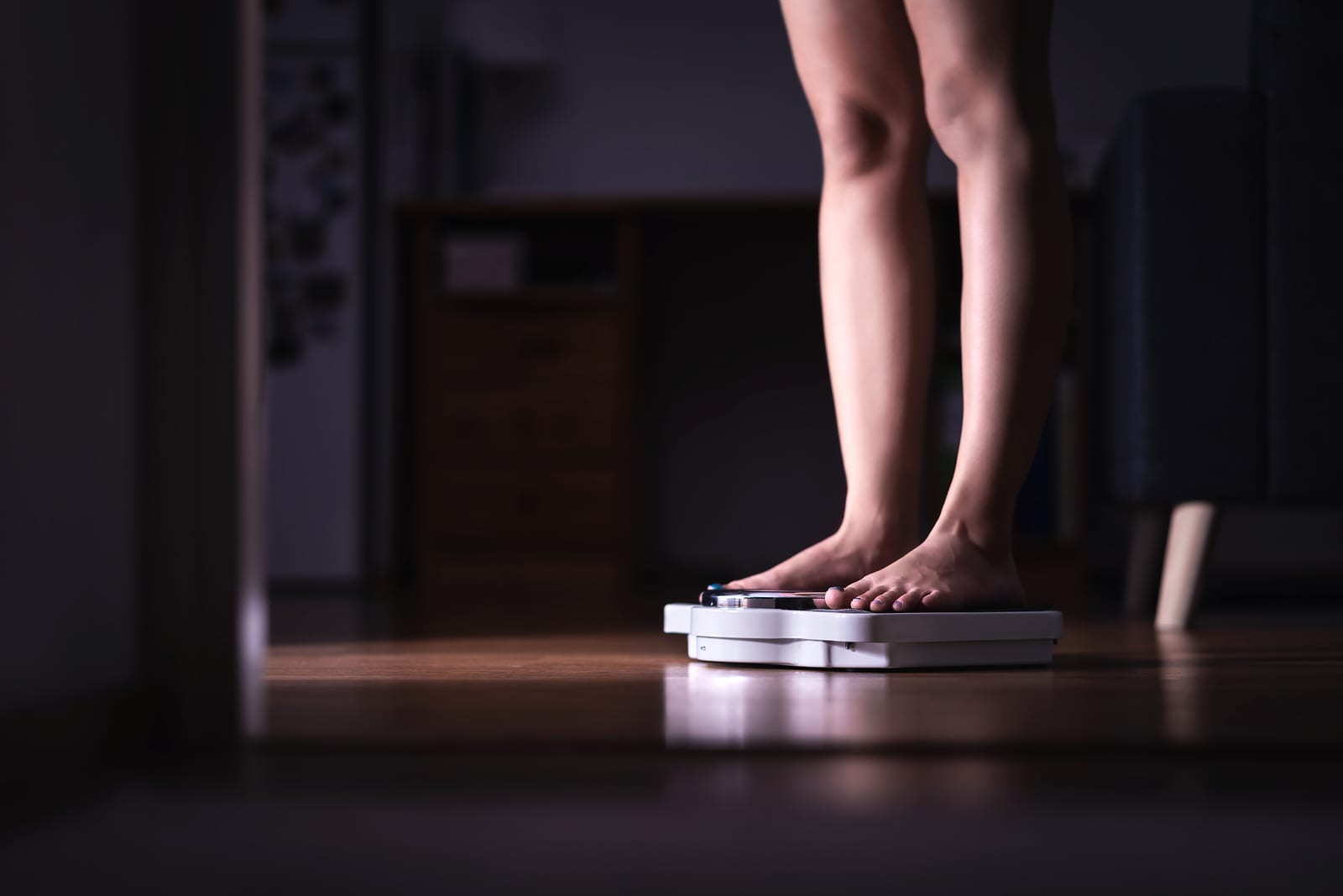 A middle aged woman stands on a scale in a dark hallway. She is checking her weight.